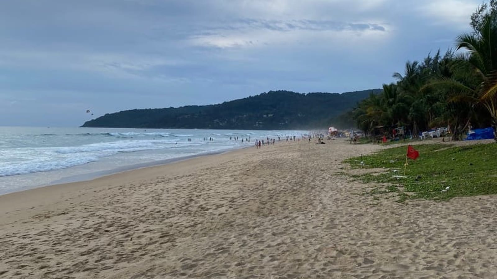 A wide beach at Karon under a grey monsoon sky, with breaking surf, a scattering of people standing in the water, and a red warning flag on the grass strip at the back of the sand