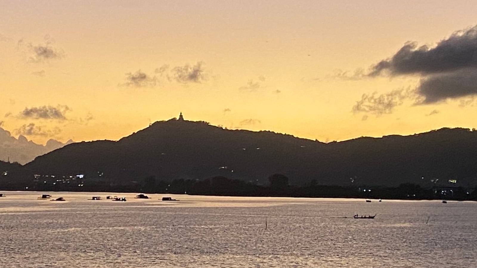 Dusk over the water on Phuket's east coast — hills in silhouette with a hilltop Buddha, longtail boats moored offshore, and the first town lights coming on